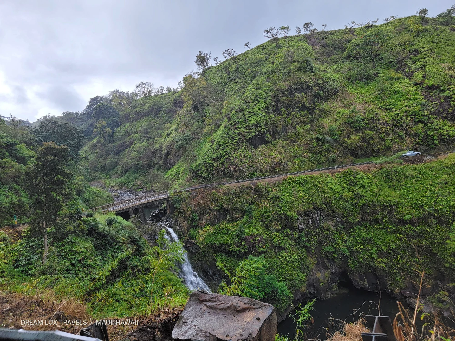 Small waterfall cascading below a bridge amid green hills along the Road to Hana.